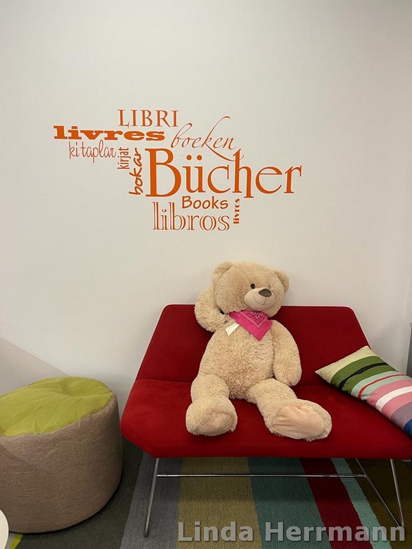Man sieht einen Plüschbären der auf einem roten Sofa sitzt. Über ihm an der Wand steht in verschiedenen Schriftarten und Größen das Wort "Bücher" in orange.