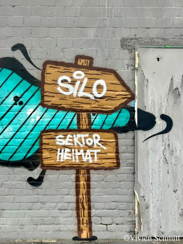 Auf diesem Bild sieht man die Außenfassade des Silodoms und eine Bemalung darauf. Es ist ein brauner Wegweiser zu sehen. Der rechte Pfeil zeigt in die Richhtung des Silodoms und das untere Schild gibt an, dass man sich gerade bei dem Gebäude des Sektor Heimats befindet.