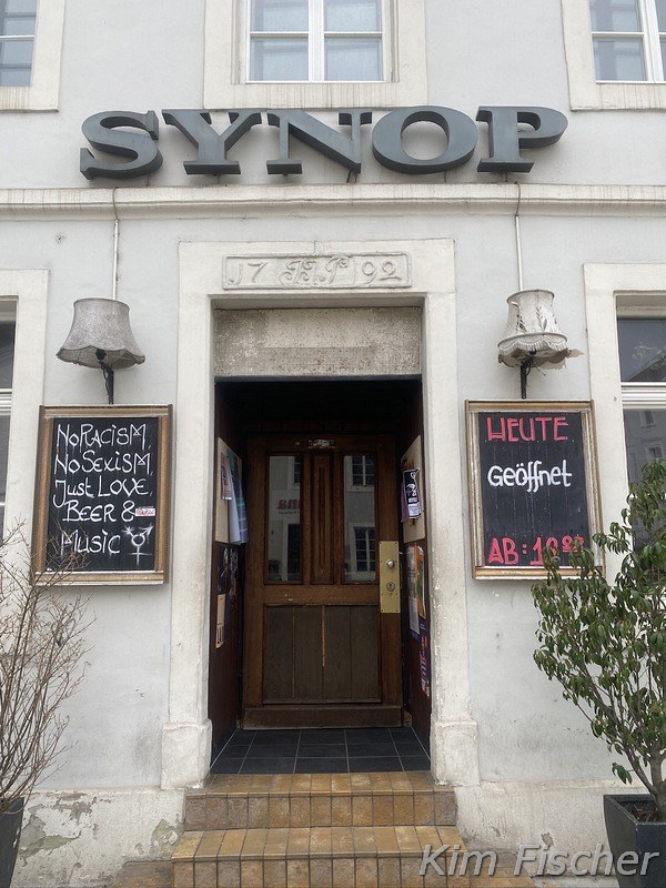 You can see the entry to the pub „Synop“. Next to the door are two lamps and to different signs. One shows the opening hours of the day and the other one a saying.