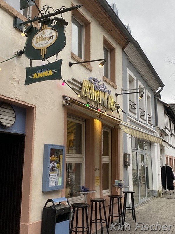 Here you can see the outdoor terrace of the pub „Tante Anna“. There are 4 chairs in front of the windows and above are a colorful fairy lights.